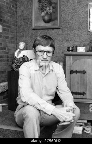 Portrait man, 00-10-1988 Whizgle News, Dutch Desk, Niederlande, 1950-2000 Ein Mann mit Brille sitzt lässig auf einem Boden, in einem hellen Hemd und einer Hose gekleidet. Er hat einen ernsthaften Ausdruck und legt seine Hände zusammen, während er von einem gemütlichen Innenraum mit einem Holzschrank, dekorativen Gegenständen und einem Blumengemälde an der Wand umgeben ist. Stockfoto