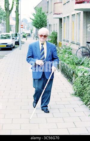 Mr. V. Hoesel blind man, 12-08-1996 Whizgle News, Dutch Desk, Niederlande, 1950-2000 ein älterer Mann mit Sonnenbrille und Blazer schlendert einen Bürgersteig hinunter und benutzt einen weißen Stock als Stütze. Er wirkt selbstbewusst und komponiert, mit ordentlich gestylten Haaren und einer gemusterten Krawatte, während Bäume und Gebäude im Hintergrund die Straße säumen. Stockfoto