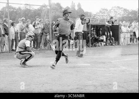 Bloemendaal - Kennemerland, MEN, Softball, 18-08-1984 Whizgle News, Dutch Desk, Niederlande, 1950-2000 Ein Baseballspieler schwingt auf dem Spielfeld, während ein aufmerksamer Fänger sich hinter der Startplatte hockt. Im Hintergrund beobachtet eine jubelnde Menge die Action, während sich die Teammitglieder in der Nähe des Dugouts versammeln. Die Szene fängt die Spannung und Intensität des Spiels ein. Stockfoto