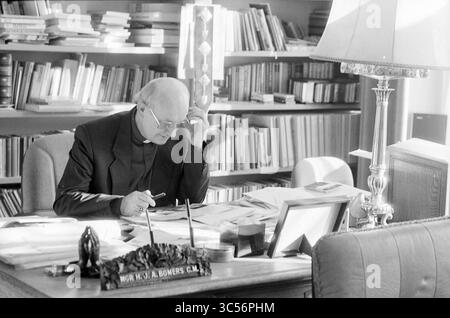 Bischof Bomers, Römisch-Katholische Kirche, 23-02-1989 Whizgle News, Dutch Desk, Niederlande, 1950-2000 Ein nachdenklicher Mann in klerikaler Kleidung sitzt an einem überfüllten Schreibtisch, umgeben von Büchern und Papieren. Er erscheint tief besinnlich, mit einem Stift in der Hand, während natürliches Licht in den Raum strömt und das organisierte Chaos seines Arbeitsplatzes unterstreicht. Stockfoto
