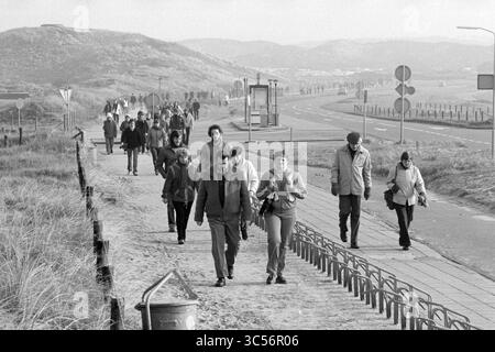 Beach Walk Haarlems Dagblad, Haarlems Dagblad etc. Und IJmuider Courant and Koeri, Walking, 31-07-1980','15-02-1981 Whizgle News, Dutch Desk, Niederlande, 1950-2000 Eine Gruppe von Menschen schlendert entlang eines Weges in der Nähe einer Straße mit sanften Hügeln im Hintergrund. Einige Menschen gehen eng zusammen, während andere verstreut sind, sich in Gespräche vertiefen oder die Umgebung genießen. Die Szene vermittelt ein Gefühl von Gemeinschaft und Freizeit, während sie den Küstenpfad entlang fahren. Stockfoto