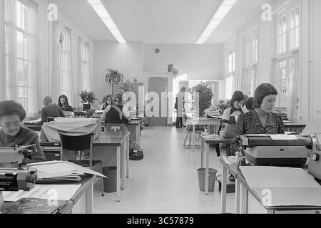 Innenraum Haarlem Town Hall, Innenraum, Haarlem Town Hall, 27-02-1975 Whizgle News, Dutch Desk, Niederlande, 1950-2000 Ein geschäftiger Bürobereich mit Schreibtischarbeiten, der sich hauptsächlich auf Schreibmaschinen konzentriert. Natürliches Licht strömt durch große Fenster und beleuchtet die organisierte Arbeitsumgebung. Mehrere Topfpflanzen sorgen für einen Hauch von Grün, während Dokumente und Schreibwaren auf jedem Arbeitsplatz übersichtlich angeordnet sind. Stockfoto