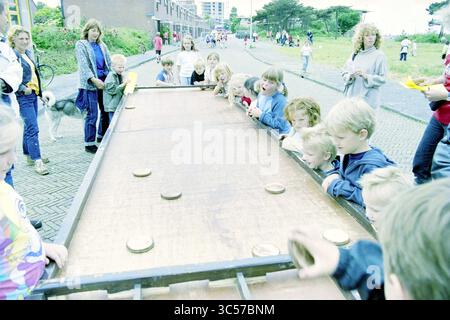 Street Play Day, Howler Buoy, IJmuiden, IJmuiden, Niederlande, 23-05-2000 Whizgle News, Dutch Desk, Niederlande, 1950-2000 Eine Gruppe von Kindern und Erwachsenen versammelt sich um einen großen Holztisch, an dem ein lebhaftes Spiel teilnimmt, auf dessen Oberfläche mehrere runde Scheiben platziert sind. Die Begeisterung erfüllt die Luft, während sich die Teilnehmer hineinlehnen und sich intensiv auf die Handlung konzentrieren. Im Hintergrund sind ein grasbewachsener Bereich und Gebäude zu sehen, die die festliche Atmosphäre verstärken. Stockfoto