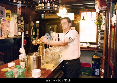 Frank Huissen, Restaurant de Ark, Haarlem, Nieuw Heiligland, Niederlande, 02-05-2000 Whizgle News, Dutch Desk, Niederlande, 1950-2000 Ein Barkeeper steht hinter einer polierten Holztheke und gießt Getränke aufmerksam aus, während Regale mit Flaschen und Gläsern den Hintergrund zieren. Das Ambiente spiegelt eine warme, einladende Pub-Atmosphäre wider, die durch eine Mischung aus Vintage-Dekor und beleuchteten Armaturen verstärkt wird. Stockfoto