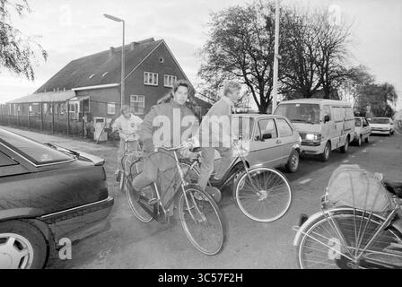 Ecke Kruisweg und Spieringweg, Radweg, Cruquius, Haarlem, Kruisweg, Niederlande, 05-11-1990 Whizgle News, Dutch Desk, Niederlande, 1950-2000 zwei Radfahrer navigieren durch eine Reihe von geparkten Autos auf einer stark befahrenen Straße. Im Hintergrund säumen ein Haus mit schrägem Dach und Bäumen die Straße und schaffen eine geschäftige urbane Szene. Stockfoto
