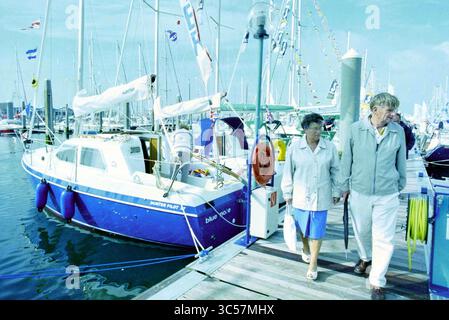 HISWA te Water, IJm., IJmuiden, Niederlande, 04-09-1995 Whizgle News, Dutch Desk, Niederlande, 1950-2000 Ein paar Spaziergänge entlang eines belebten Yachthafens mit einem blauen Segelboot in der Nähe. Verschiedene Fahnen flattern in der Brise und verleihen der Szene eine lebendige Atmosphäre, während sie ihren Tag am Wasser genießen. Stockfoto