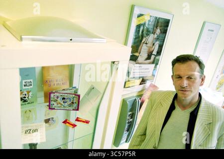 Allgemeinarzt Venserpolder, Amsterdam, Niederlande, 03-05-2001 Whizgle News, Dutch Desk, Niederlande, 1950-2000 Ein Mann in einem hellen Blazer steht neben einer Vitrine mit verschiedenen Gegenständen, darunter bunten Karten und Broschüren, vor dem Hintergrund von informativen Postern an der Wand. Stockfoto
