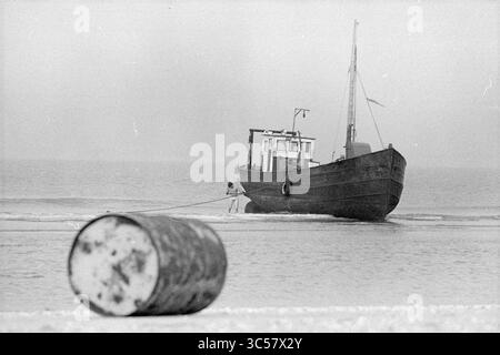 Stranded Ship, Parnassia Beach, Schiffe, 16-10-1974 Whizgle News, Dutch Desk, Niederlande, 1950-2000 Ein verwittertes Fischereifahrzeug liegt in der Nähe des Ufers vor Anker, mit Seilen, die sich bis zum Strand erstrecken, wo ein rostiges Fass auf dem Sand ruht, was einen starken Kontrast zur ruhigen Meereskulisse bildet. Stockfoto