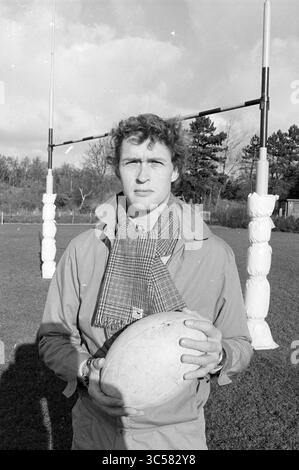 Peperkamp Rugby Coach, Rugby, 14-11-1981 Whizgle News, Dutch Desk, Niederlande, 1950-2000 Ein junger Mann steht selbstbewusst auf einem Rugbyfeld, hält einen Rugbyball und trägt einen stylischen Schal. Hinter ihm bilden Torpfosten den Rahmen für die Szene, vor einem Hintergrund von Bäumen und einem bewölkten Himmel. Stockfoto
