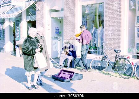 Schönes Wetter, Grote Houtstraat, Haarlem, Grote Houtstraat, Niederlande, 25-02-2000 Whizgle News, Dutch Desk, Niederlande, 1950-2000 Eine Straßenszene zeigt zwei elegant gekleidete ältere Personen, die an einem Musiker, der Gitarre spielt, und einem anderen Mann, der Geige spielt, vorbeilaufen. In der Nähe neigt sich ein Fahrrad an ein Gebäude, und ein Gitarrenkoffer liegt offen auf dem Boden, was einen Moment künstlerischen Ausdrucks in einer lebendigen Umgebung im Freien suggeriert. Stockfoto