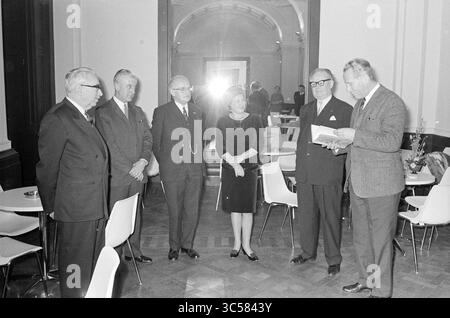 Jubilarian erhält Glückwünsche und ein Buch Geschenk Whizgle News, Dutch Desk, Niederlande, 1950-2000 Eine Gruppe von sechs Personen steht in einem stilvollen Innenraum, in Gesprächen. Sie sind in formeller Kleidung gekleidet, zwei Männer lesen von einem Blatt Papier, während andere aufmerksam zuhören. Das Ambiente bietet moderne Sitzmöbel und minimalistische Einrichtung, die eine elegante Atmosphäre schaffen. Stockfoto