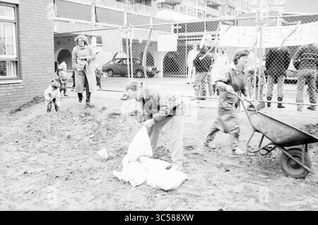 Squatting Spielfeld Engelenburg Schalkwijk, Squatting, Play and Toys, Haarlem, Engelenburg, Niederlande, 29-03-1984 Whizgle News, Dutch Desk, Niederlande, 1950-2000 Eine Gruppe von Einzelpersonen, darunter Kinder und Erwachsene, arbeitet in der Gemeinde, bereitet einen Sandgrundstück mit Taschen und Schubkarre vor, während andere hinter einem mit Bannern geschmückten Zaun beobachten. Stockfoto