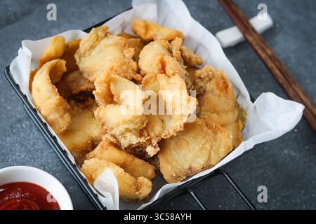 Goldbraune, knusprige Fischstücke, serviert in einem stilvollen Korb aus schwarzem Draht. Eine kleine rote Schüssel Soße ist im Hintergrund zu sehen. Stockfoto