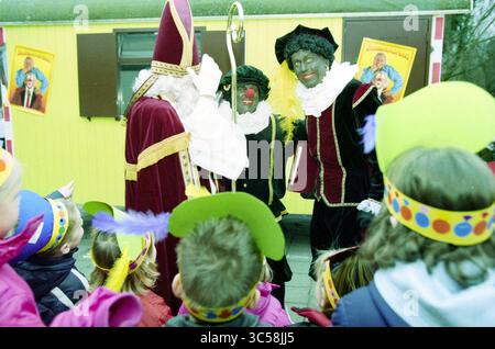 Saint Nicholas on the Viersprong, 04.12-2002 Whizgle News, Dutch Desk, Niederlande, 1950-2000 Ein festliches Treffen mit einer Gruppe von Charakteren in aufwendigen Kostümen, darunter eine Figur in Bischofskleidung und zwei Figuren in traditioneller Kleidung, die alle mit aufgeregten Kindern in bunten Hüten mit Federn und Polka Dots zu tun haben. Helle, fröhliche Dekorationen sorgen für eine verspielte Szene. Stockfoto