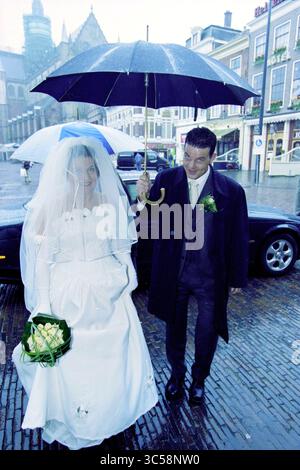Hart en Schrama Wedding, Haarlem, Niederlande, 22-03-2001 Whizgle News, Dutch Desk, Niederlande, 1950-2000 Ein frisch vermähltes Paar geht Hand in Hand durch eine regnerische Straße, die Braut in einem schönen weißen Kleid und Schleier, hält einen Blumenstrauß, während der Bräutigam sie beide mit einem Regenschirm abschirmt. Die charmante Umgebung bietet historische Gebäude im Hintergrund. Stockfoto