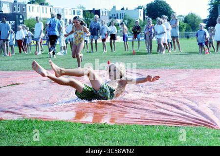 Folien üben, Kinheim Youth, 18-08-1998 Whizgle News, Dutch Desk, Niederlande, 1950-2000 Ein Kind plätschert fröhlich auf einer nassen Rutsche, umgeben von jubelnden Kollegen und genießt einen sonnigen Tag voller Spaß und Spiele auf dem Gras. Stockfoto