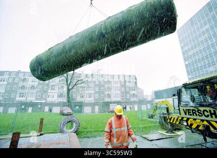 Entsorgung des alten Öltanks von der Hochschule Amsterdam, Amsterdam, Niederlande, 14-12-1999 Whizgle News, Dutch Desk, Niederlande, 1950-2000 Ein Bauarbeiter, der einen Schutzhelm und eine leuchtend orange Jacke trägt, wird mit einem Kran über eine Baustelle gehoben, wobei Gebäude im Hintergrund sichtbar sind und Schneeflocken um sie herum fallen. Stockfoto