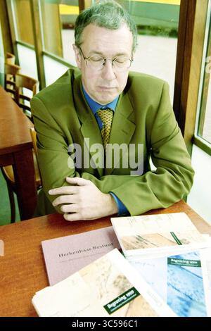Mr. Verhoef, Sloterdijk, Sloterdijk, 24-10-1999 Whizgle News, Dutch Desk, Niederlande, 1950-2000 Ein Mann im Anzug sitzt mit geschlossenen Augen und einem nachdenklichen Ausdruck an einem Tisch. Verschiedene Dokumente und Berichte sind vor ihm verteilt, die eine Atmosphäre der Nachdenklichkeit oder der Konzentration auf die Arbeit suggerieren. Die Lage scheint ein ruhiger Raum mit Holzmöbeln und einem Fenster im Hintergrund zu sein. Stockfoto