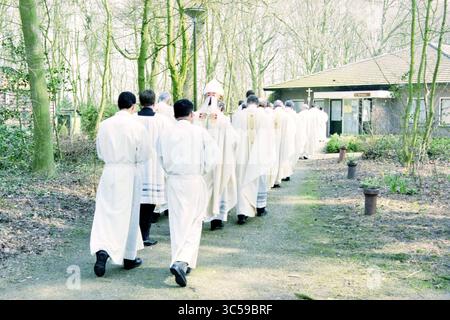 Ordination von vier Diakonen, Heiloo, Heiloo, 24-03-1999 Whizgle News, niederländisches Büro, Niederlande, 1950-2000 Eine Gruppe von Individuen, die in weißen Gewändern gekleidet sind, geht auf einem Waldweg zu einem rustikalen Gebäude inmitten der Bäume. Stockfoto