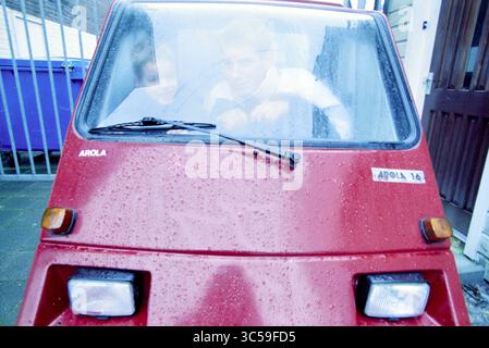 Mann und Sohn in der Automarke Arola, 00-02-2001 Whizgle News, Dutch Desk, Niederlande, 1950-2000 Ein knallrotes dreirädriges Fahrzeug, das in einer engen Gasse geparkt ist, mit zwei Personen im Inneren, die aufmerksam durch die Windschutzscheibe blicken, wobei sich Regentropfen an der Scheibe anklammern. Die Umgebung weckt ein Gefühl der Neugier und Verspieltheit. Stockfoto