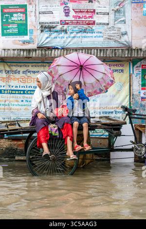 Mutter und Kind unter Regenschirm auf einer Rikscha in der überfluteten Straße. Die Abholzung von Wasserfällen ist in Bangladesch ein ernstes Problem, insbesondere während des Monsuns Stockfoto