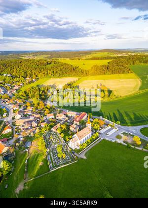 Bohutin Town ist von oben erfasst und zeigt sein bezauberndes Layout. Grüne Felder erstrecken sich in die Ferne, ergänzt durch eine historische Kirche und einen Friedhof. Die Sonne wirft lange Schatten. Stockfoto