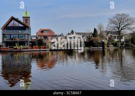 In Ouderkerk aan de Amstel befinden sich Häuser am Wasser, die sich in einem ruhigen Fluss unter einem hellen Himmel spiegeln und die bezaubernde niederländische Dorfszene einfangen. Stockfoto