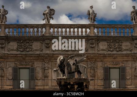 Klassische Marmorstatuen von sechs römischen Gottheiten, Apollo, Jupiter, Venus, Quecksilber, Minerva und Herkules, Linie barocke Balustrade um 1626 in den Palazzo Maffei auf der Piazza delle Erbe, Verona, Veneto, Italien. Unter der Säule aus dem 16. Jahrhundert steht der geflügelte Markuslöwe, das Symbol von Venedig und der Republik Venedig. Die Inschrift auf dem offenen Buch übersetzt: „Friede zu dir, Mark, mein Evangelist“. Stockfoto
