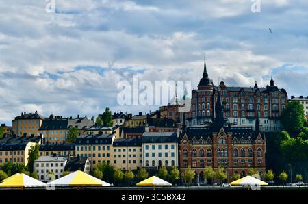 Malerische Stadtlandschaft von Södermalm in Stockholm, Schweden, mit historischer Architektur unter bewölktem Himmel. Farbenfrohe Gebäude und gelbe Sonnenschirme. Stockfoto