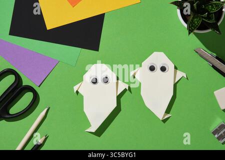 Zwei handgemachte Origami-Geister aus weißem Papier mit guten Augen sind auf einem grünen Hintergrund angeordnet, umgeben von bunten Blättern, Bleistiften, Scheren, Cutter, Stockfoto
