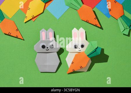 Zwei Origami-Häschen, eines grau und eines weiß, mit rosa Ohren und guten Augen, sitzen auf leuchtendem grünem Papier, umgeben von Origami-Karotten Stockfoto