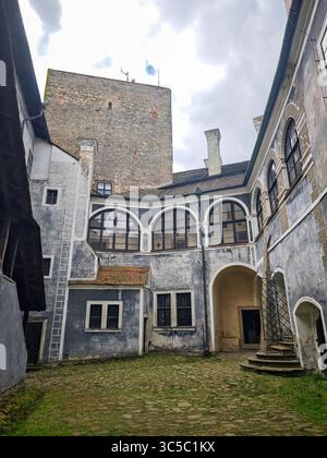 Innenhof der Burg Buchlov in der Tschechischen Republik, mit verwitterten Steinmauern, Bogenfenstern und einem historischen Turm unter bewölktem Himmel. Stockfoto