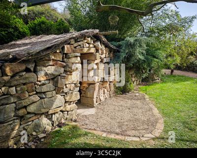 Traditioneller Trockensteinunterstand mit einem Strohdach in einer Gartenanlage. Die Struktur besteht aus gestapelten Natursteinen und ist von Grün und umgeben Stockfoto