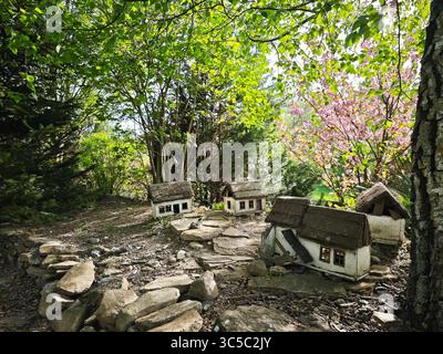 Rustikale Miniatur-Cottages mit Strohdach und Fliesendächern in einem Waldgarten, umgeben von Frühlingsgrün und blühenden rosa Blumen. Stockfoto