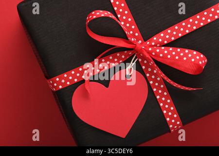Karte mit schwarzer Geschenkbox mit leerem herzförmigem Etikett auf rotem Hintergrund. Platz für Ihren Text Stockfoto
