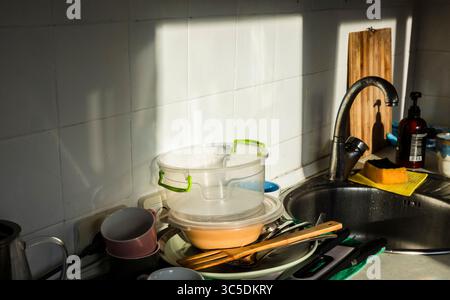 Schmutziges Geschirr, Besteck, Tassen und Plastikbehälter, die ein Spülbecken überladen, mit Sonnenlicht, das Schatten auf weiße Fliesen wirft, schaffen eine unordentliche Szene Stockfoto