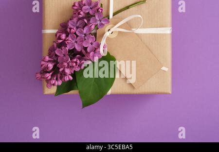 Braune handwerkliche Geschenkbox mit leerem Tag, lila Blumen auf violettem Hintergrund. Platzieren für Ihren Text Stockfoto