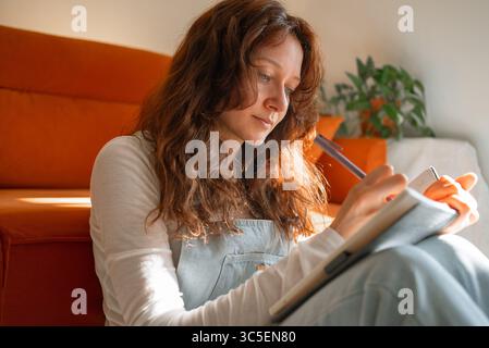 Konzentrierte junge Frau, die Notizen auf Notizbuch schreibt, sitzt zu Hause auf dem Boden, trägt legere Kleidung, mit einer orangefarbenen Couch im Hintergrund Stockfoto