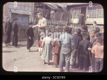U.S. Marine Staff Sergeant verteilt Süßigkeiten an koreanische Waisenkinder außerhalb eines lokalen Waisenhauses in Masan, Korea, April 1957. Kinder kommen in der Schlange zusammen, während ältere Schüler in der Nähe einer Tafel mit handgeschriebenem koreanischem Schriftzug beobachten. Teil der humanitären Hilfsmaßnahmen der USA nach dem Koreakrieg. Stockfoto