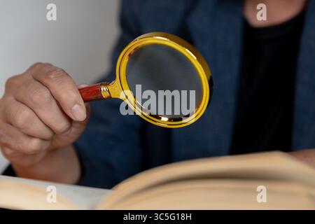 Ein älterer Mann, der mit einer Lupe ein Buch liest, das Sehschwierigkeiten und das Altern des Sehvermögens veranschaulicht. Stockfoto