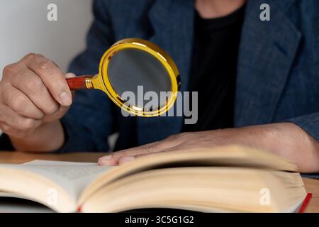 Ein älterer Mann, der mit einer Lupe ein Buch liest, das Sehschwierigkeiten und das Altern des Sehvermögens veranschaulicht. Stockfoto