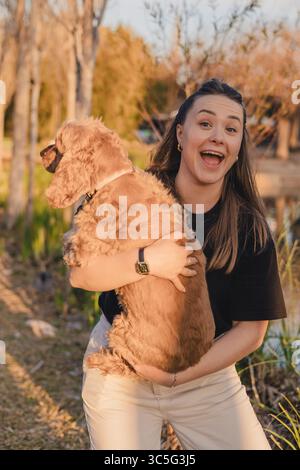 Eine junge kaukasische Frau mit langen braunen Haaren lächelt, während sie einen goldenen Cocker Spaniel in einer natürlichen Umgebung mit Bäumen und Gras hält. Stockfoto