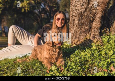 Eine junge kaukasische Frau mit langen braunen Haaren sitzt neben einem Baum. Ein goldener Cocker Spaniel liegt auf dem Gras und genießt den sonnigen Tag. Stockfoto