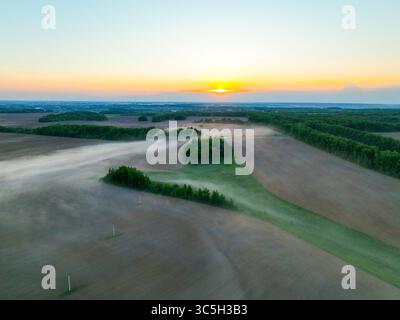 Ein Blick aus der Vogelperspektive fängt Nebel ein, der bei Sonnenaufgang über bewirtschafteten Feldern und Waldflächen wirbelt und eine ruhige ländliche Landschaft präsentiert Stockfoto