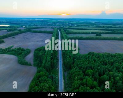 Eine Rundfahrt aus der Vogelperspektive zeigt eine lange Straße, die von dichten grünen Wäldern flankiert wird und sich durch weite Felder unter einem Sonnenuntergang erstreckt Stockfoto