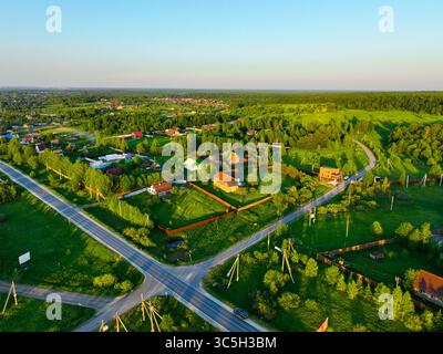 Ein Blick aus der Vogelperspektive bietet eine ländliche Straßenkreuzung, umgeben von verstreuten Häusern und einer üppigen grünen Landschaft unter dem warmen Licht des Sonnenuntergangs Stockfoto