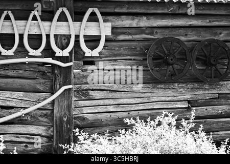 Schwarz-weiß-Foto einer traditionellen hölzernen Scheunenwand mit alten Wagenrädern und hängendem Pferdejoch. Alte ländliche Architektur und Landwirtschaftsgeräte o Stockfoto