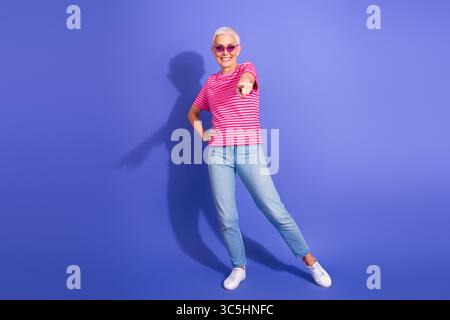 Elegante, reife Frau, die selbstbewusst in einem stilvollen Outfit vor lila Hintergrund Gesten für Lifestyle- und Modeinhalte verleiht Stockfoto