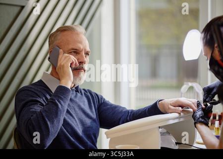 Kaukasier mittleren Alters, der mit dem Smartphone spricht, während er Maniküre von einer weiblichen Nageltechnikerin im Salon erhält, am Tisch neben dem Fenster sitzt und fokussiert Stockfoto