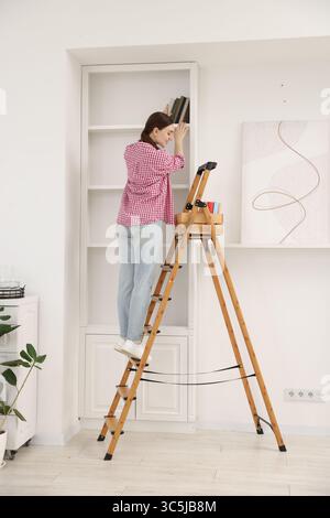 Frau, die zu Hause Bücher in Regale legt, indem sie Leiter benutzt Stockfoto